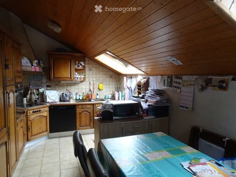 A cozy kitchen with wooden cabinets, a stove, dishwasher, microwave, refrigerator, and a dining table with chairs. The room features a slanted wooden ceiling and a skylight window.