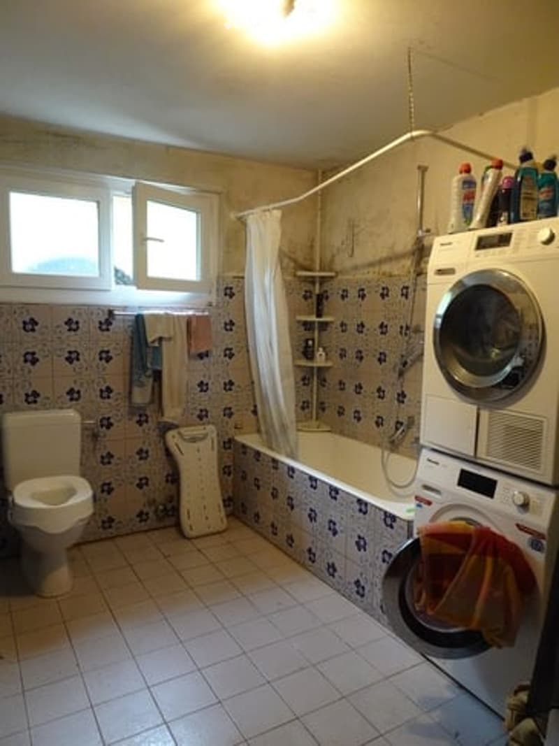 Bathroom with tiled walls and floor, toilet, bathtub, shower curtain, washing machine, and cleaning supplies on a shelf.
