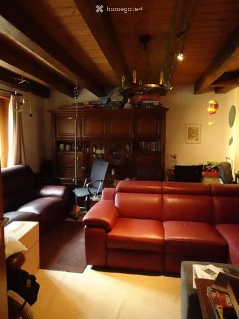 Living room with exposed wooden beams, large red leather sofa, dark brown leather armchair, wooden cabinet, and a hanging chandelier.