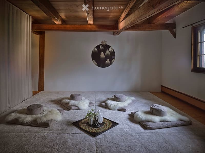 Minimalist bedroom with wooden beams, meditation cushions, furs, and a small table with a vase and flower.