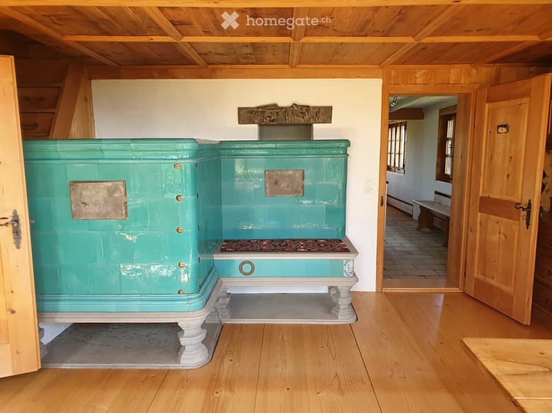Room with wooden floors and ceiling, featuring a large teal fireplace with decorative metal plates and a small bench visible through an open doorway.