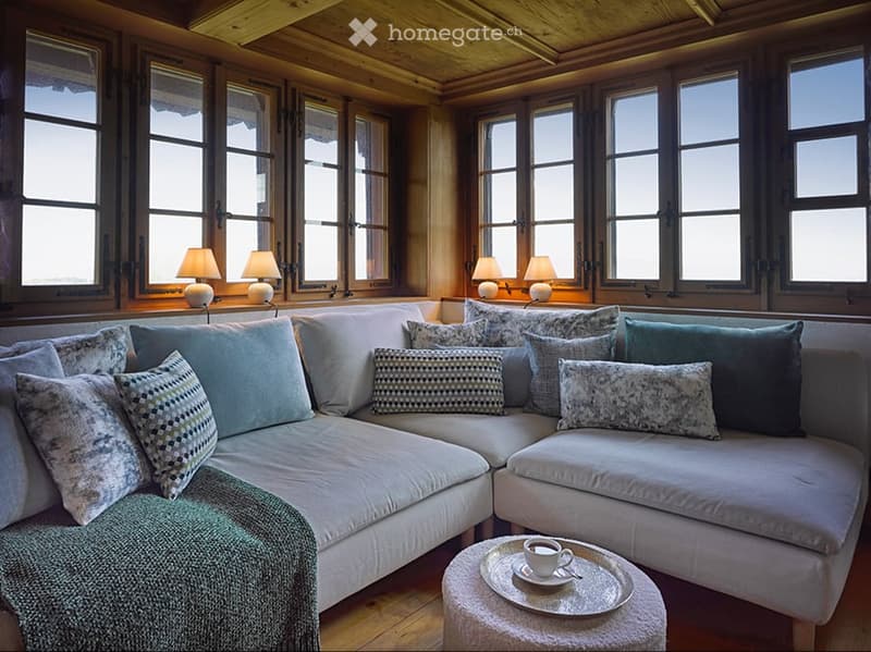 Cozy living room with large windows, wooden ceiling, and a sectional sofa adorned with multiple pillows. A small coffee table holds a cup and saucer.