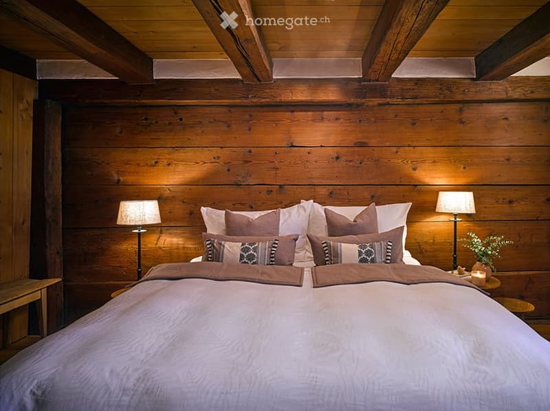 Cozy bedroom with wooden walls and ceiling, large bed with multiple pillows and blankets, nightstands with lamps, small table with candle and plant.