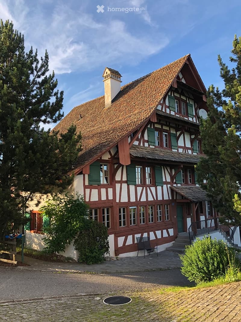 Traditional half-timbered house with a steeply pitched roof, green shutters, and a chimney. The building is surrounded by trees and has a small porch with a bench.