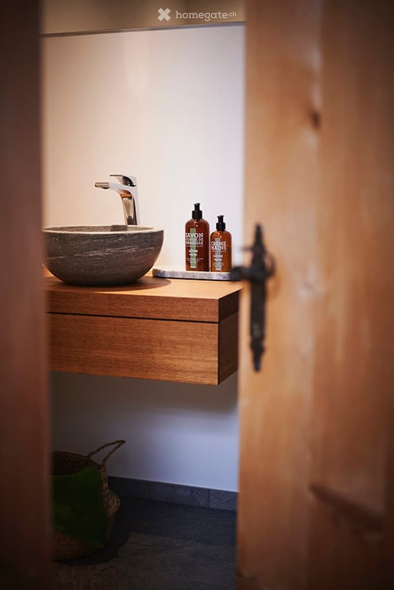 Modern bathroom with a wooden sink cabinet, stone sink bowl, sleek faucet, and decorative bottles on the counter. A wicker basket is placed on the floor.