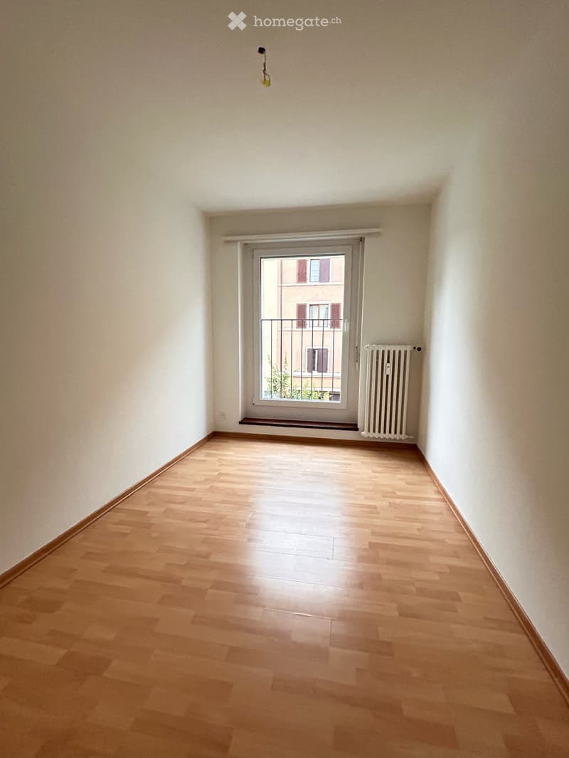 Empty room with wooden flooring, white walls, a window with a metal security grate, and a radiator on the wall.