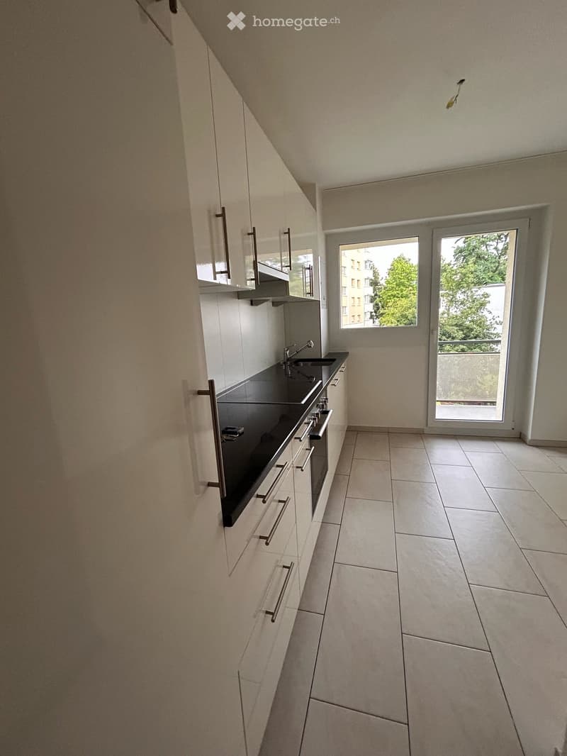 Modern kitchen with white cabinets, black countertop, stainless steel sink, and built-in oven. Features large windows and a sliding glass door leading to a balcony.