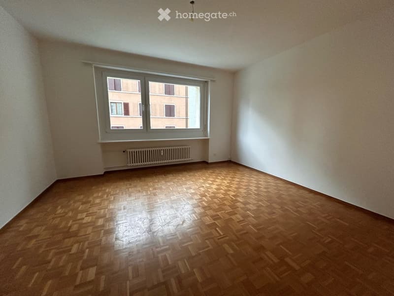 Empty room with wooden parquet flooring, white walls, and a large window with a radiator below it.