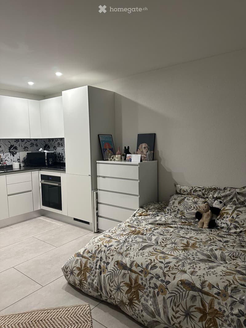 Modern bedroom with a large bed, nightstand, and adjacent kitchen area with white cabinets and appliances.