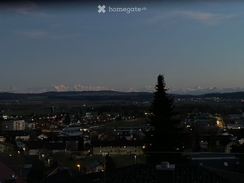 Nighttime view of a town with scattered lights, houses, and distant mountains under a dark sky.