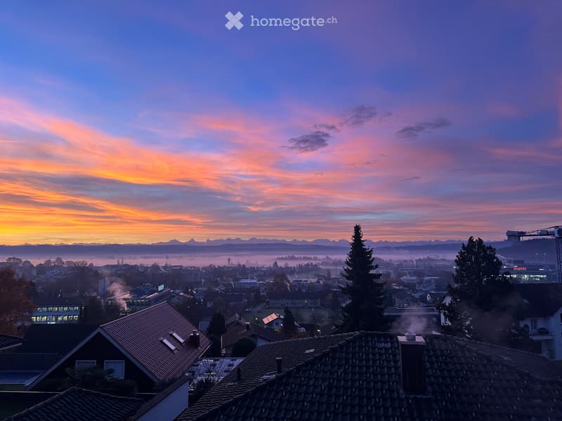 Panoramic view of a cityscape at sunset, featuring rooftops, distant mountains, and a colorful sky.