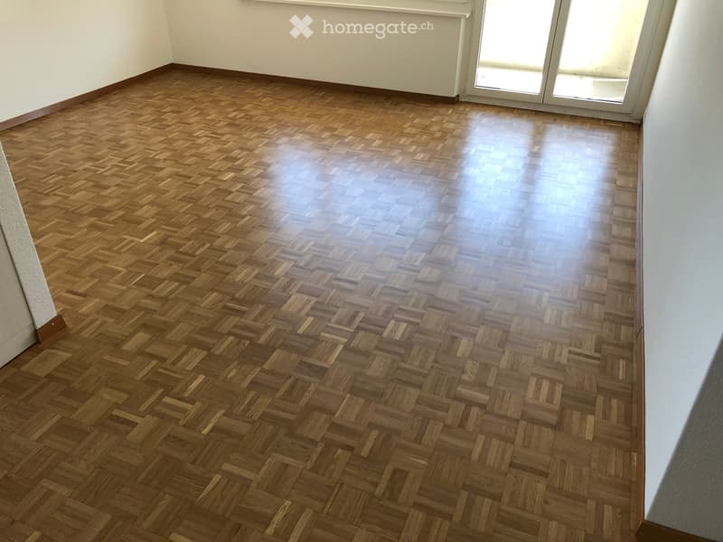 Empty room with herringbone-patterned wooden flooring and white walls, featuring a large sliding glass door at the far end.