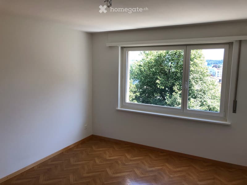 Empty room with wooden flooring and a large window showing greenery outside.