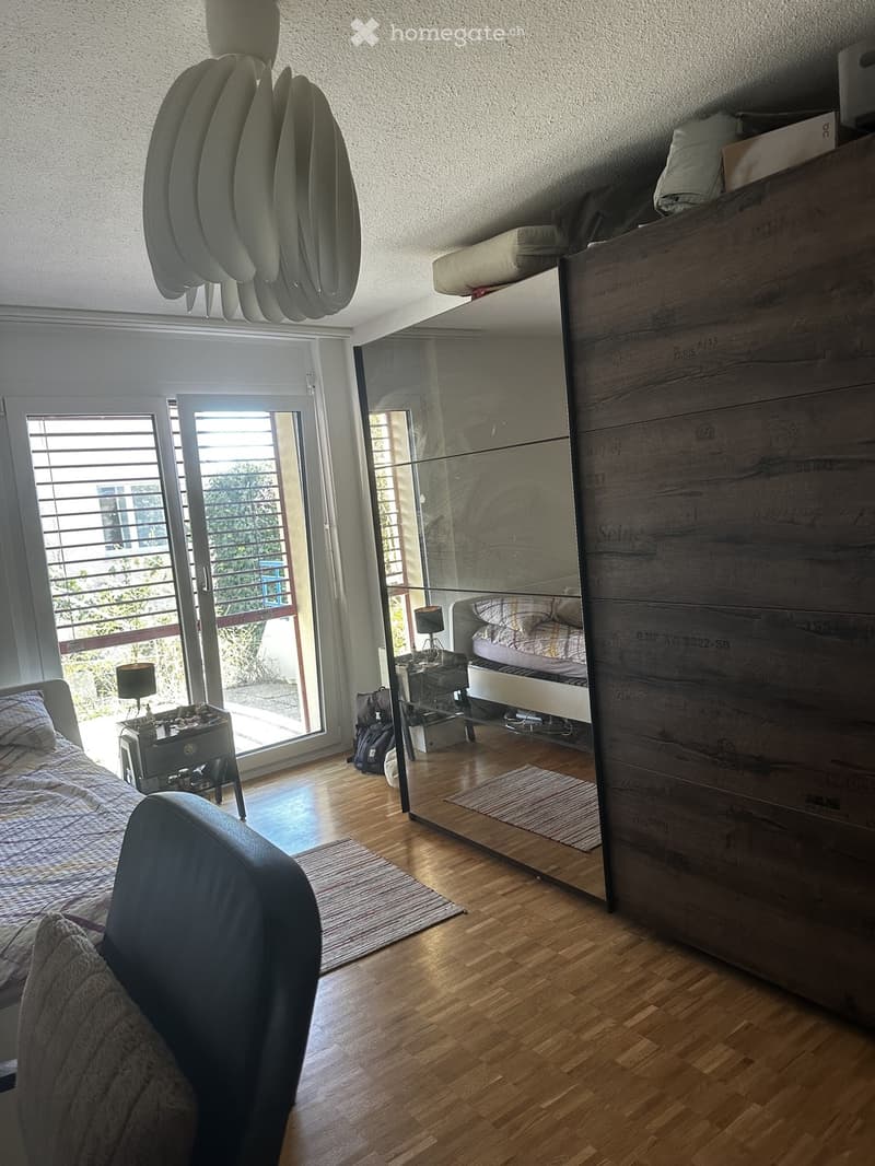 Modern bedroom with wooden flooring, large bed, nightstand, chair, wardrobe, and a mirrored closet. Large sliding glass door leads to a balcony.