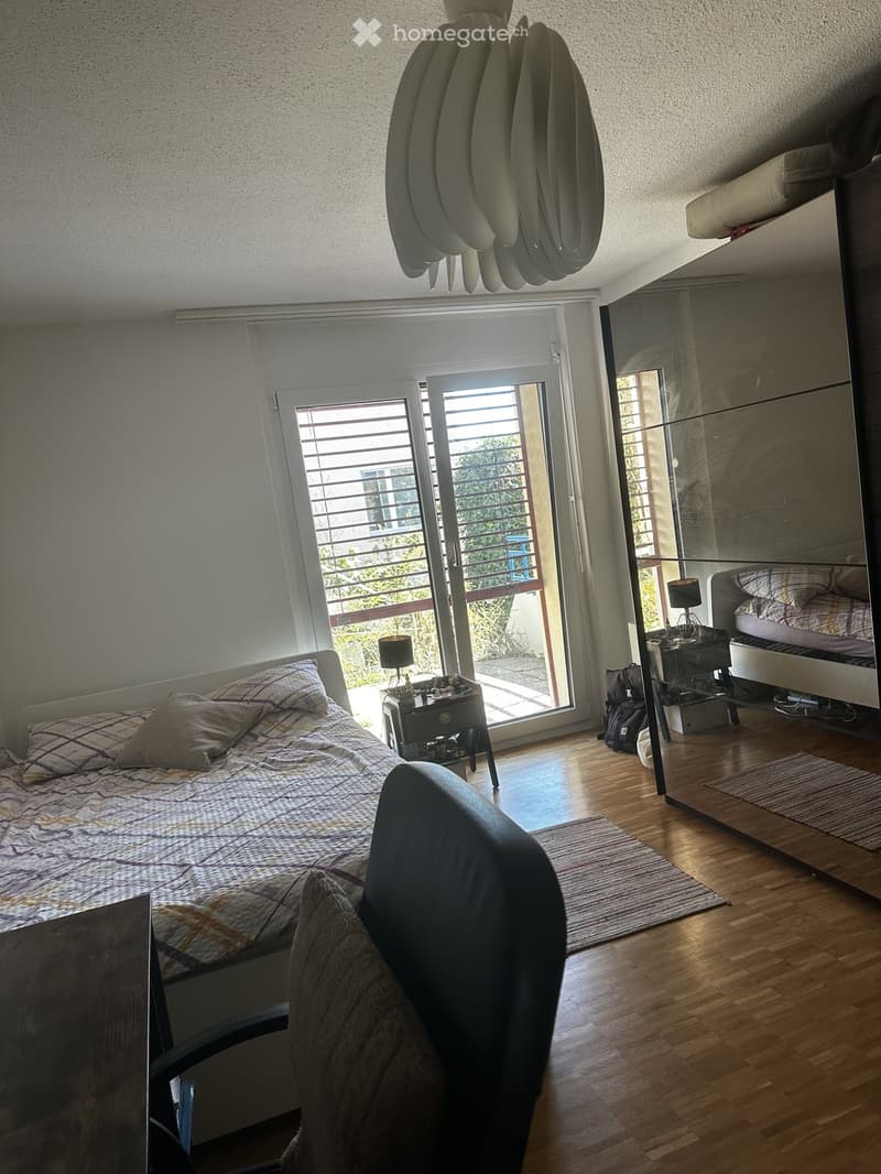 Bright bedroom with wooden flooring, large window with blinds leading to a balcony, bed with patterned bedding, nightstand with lamp, chair, and mirror.