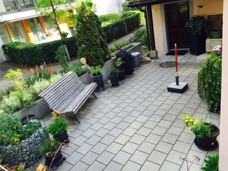 A well-maintained garden area with a tiled patio, potted plants, a wooden bench, and a small statue. The space is bordered by neatly trimmed hedges and features a variety of flowers and greenery.