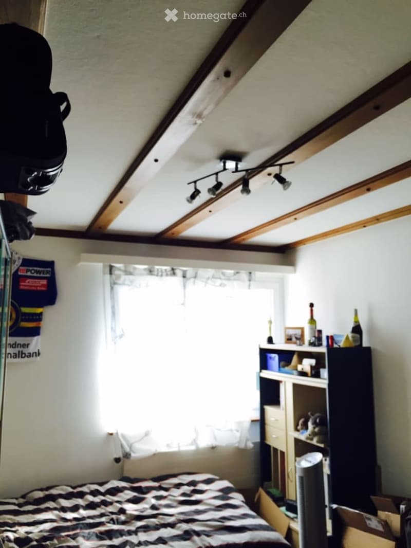 A cozy attic bedroom with exposed wooden beams, a slanted ceiling, and modern track lighting. The room features a neatly made bed, a wardrobe, a shelf with various items, and a small fan on the floor.