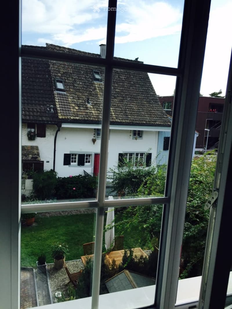 View from a window showing a garden area with a wooden table, chairs, potted plants, and a neighboring house with a tiled roof and white facade.