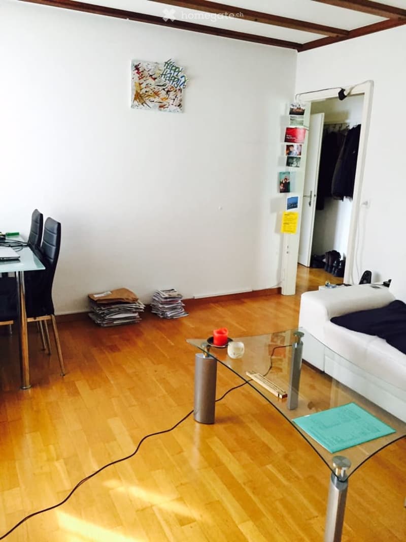Living room with wooden floor, white walls, a white sofa, a glass coffee table, a dining table with chairs, and a mirror on the wall.