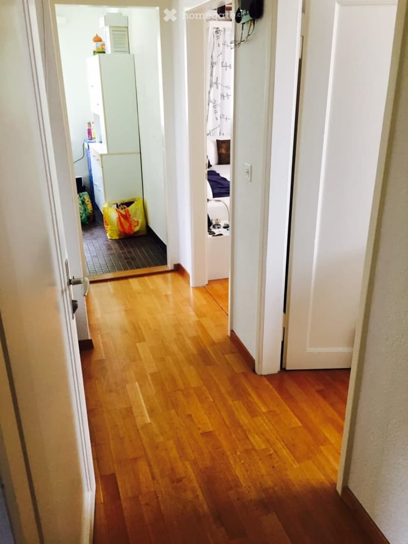 A narrow hallway with wooden flooring and white walls, leading to a bedroom and a kitchen area.