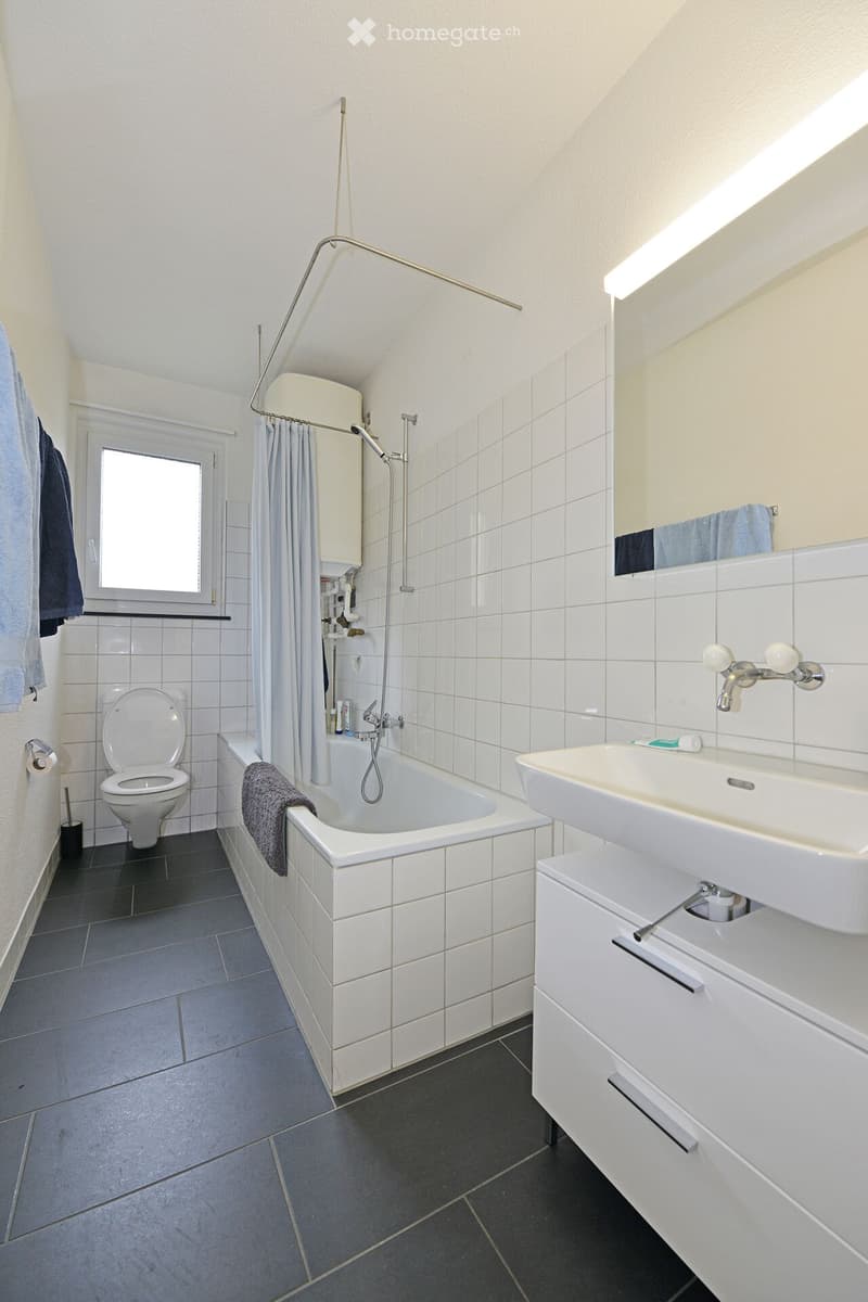 Modern bathroom with white tiles, a bathtub, toilet, sink, and a large mirror. Features a shower curtain, towels, and a window for natural light.