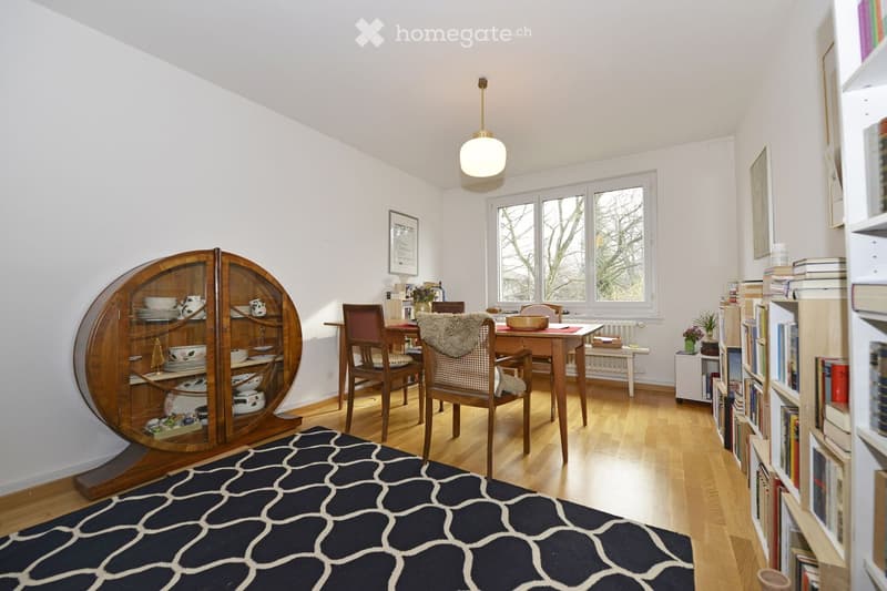 Brightly lit room with wooden flooring, a round glass-fronted cabinet, a dining table with chairs, a bookshelf filled with books, and a patterned rug.