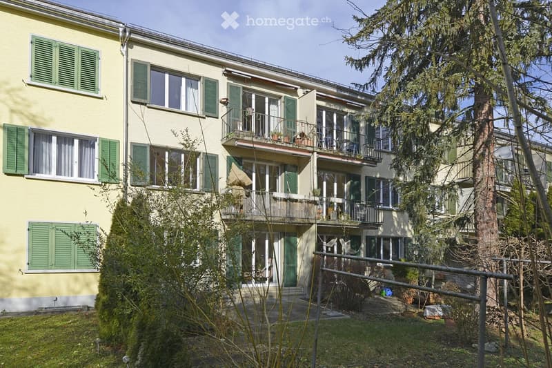 A three-story residential building with yellow walls, green shutters, and multiple balconies. The building features large windows and is surrounded by trees and greenery.