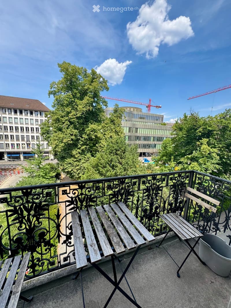 Balcony with ornate black metal railing, wooden benches, and a view of trees and buildings.