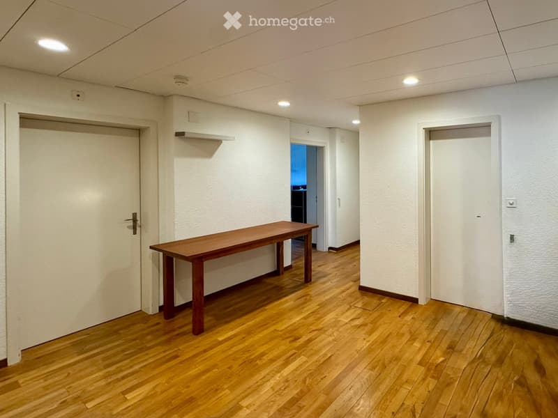 A small, unfurnished room with wooden flooring, white walls, and a single wooden table placed against the wall. Two doors are visible, one leading to another room.