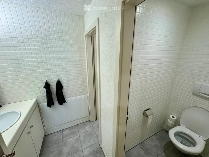 A small bathroom with white tiled walls and floor, featuring a sink with a cabinet, a toilet, and two black towels hanging on the wall.