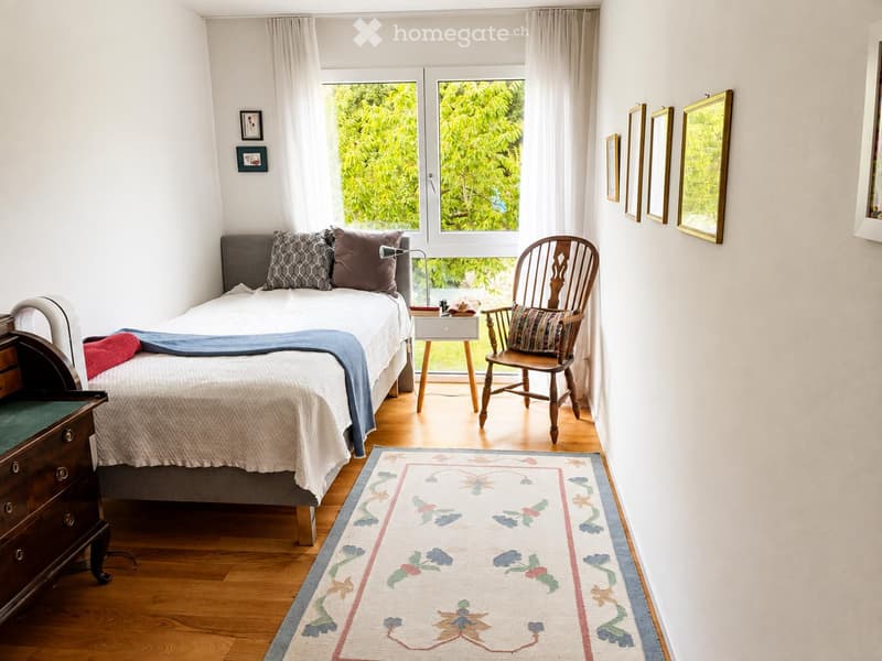 Cozy bedroom with a single bed, wooden chair, nightstand, lamp, dresser, and a decorative rug. Large windows with sheer curtains let in natural light.