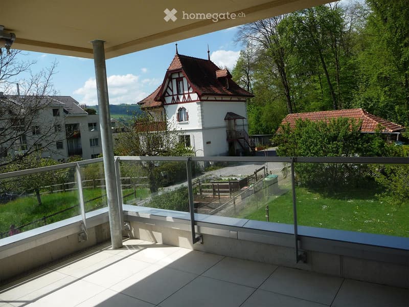 Balcony with glass railing, tiled floor, overlooking a landscaped area with a small table, chairs, and a view of a historic building with a red-tiled roof and white walls.