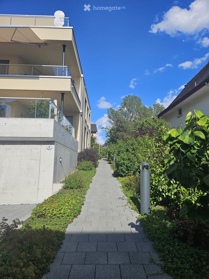 A paved pathway lined with greenery leads to a modern building with balconies and a satellite dish on the roof.