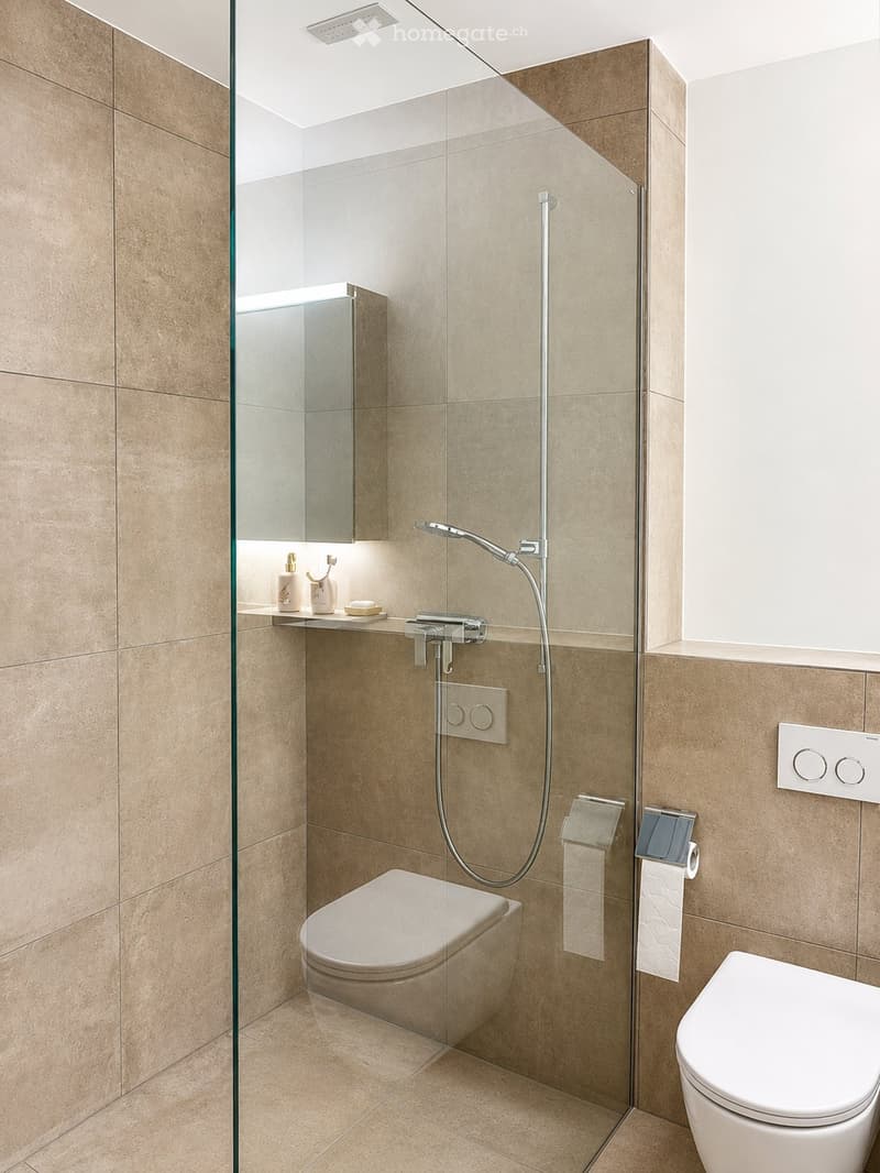 Modern bathroom with beige tiles, glass shower enclosure, two toilets, a handheld showerhead, and a small shelf holding toiletries.