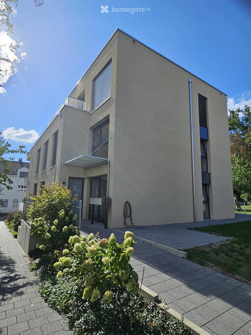 Modern two-story building with a flat roof, large windows, and a small balcony on the upper floor. The facade is light-colored with clean lines, and the entrance features a covered walkway. The surrounding area includes a paved walkway and some greenery.