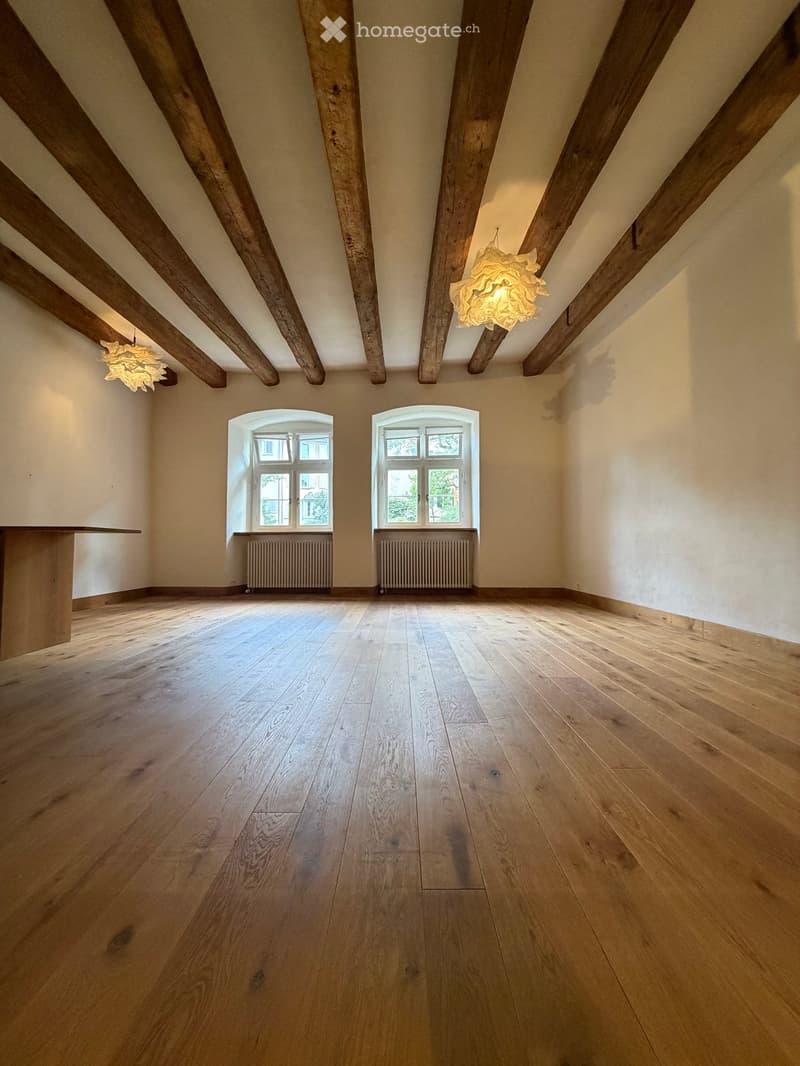 Empty room with wooden floor, exposed wooden beams, two windows, and two hanging lamps.