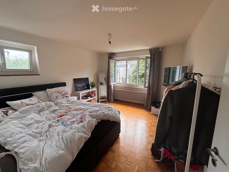 Cozy bedroom with wooden flooring, large window with view, bed with decorative pillows, nightstand, mirror, and clothing rack.