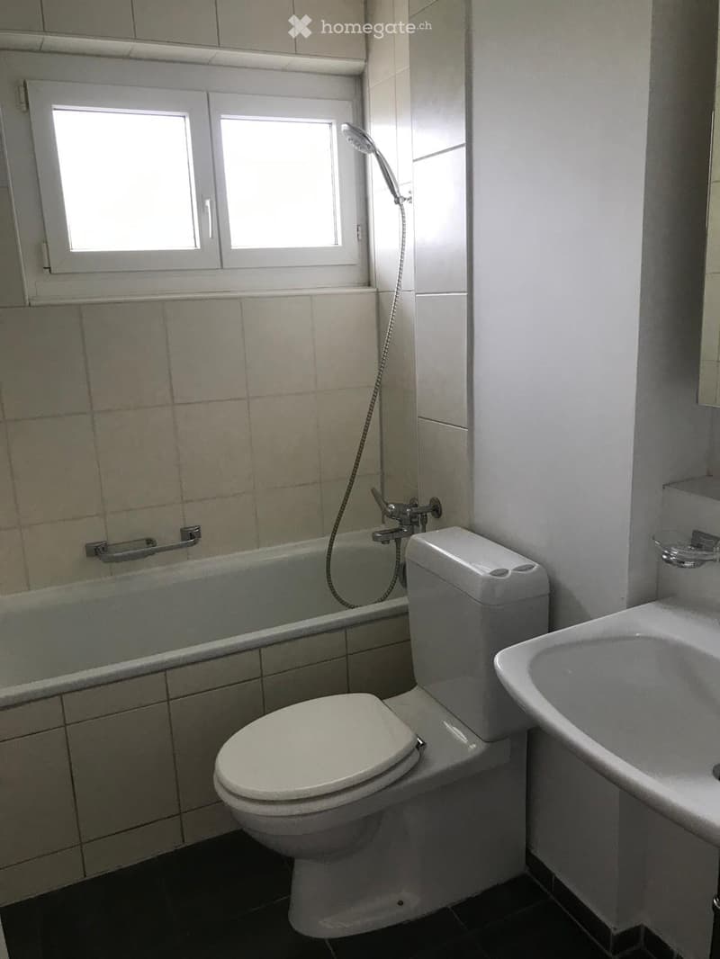 Bathroom with a toilet, bathtub, sink, and showerhead. The walls are tiled, and there is a window above the bathtub.