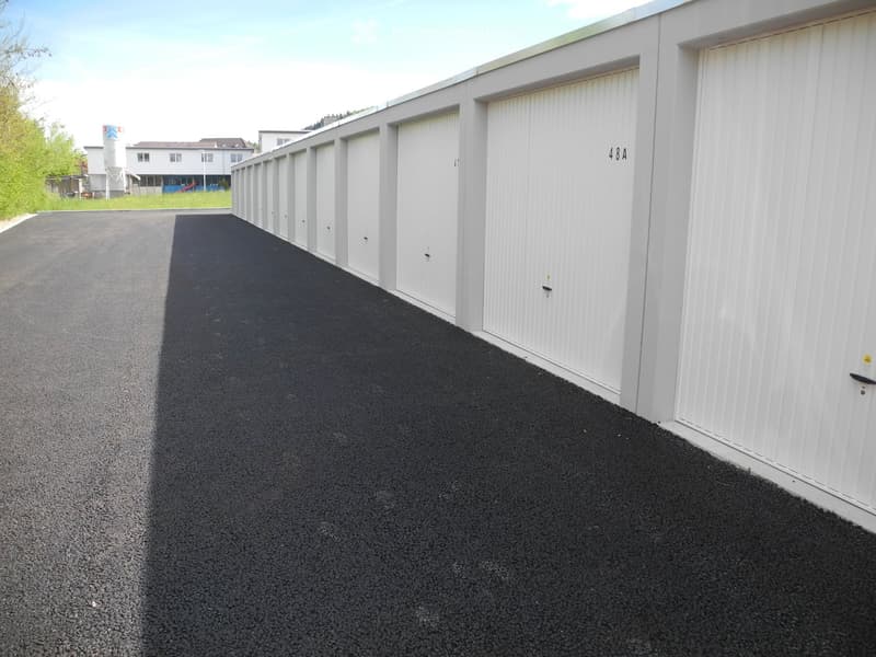 A row of white garage units with closed doors, numbered "484" on the right, situated on a paved asphalt area with a grassy area and distant buildings in the background.