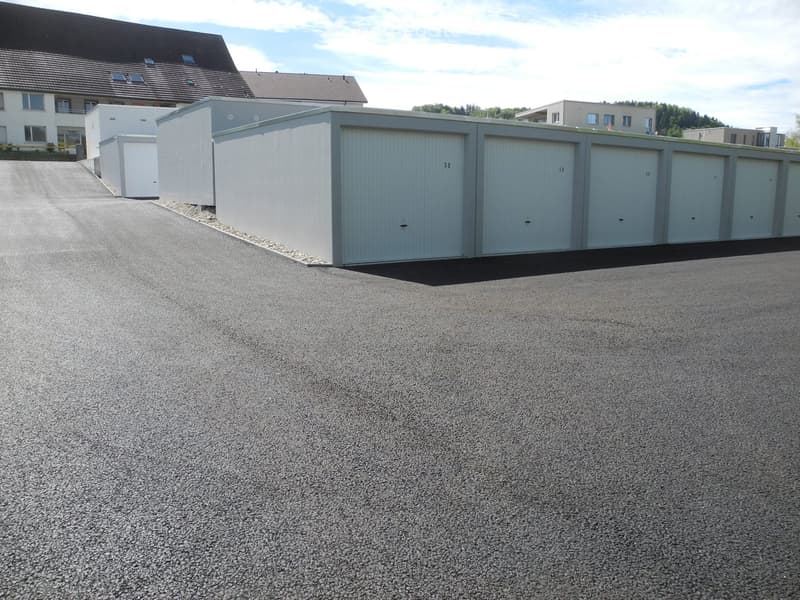 A row of grey storage units with closed doors, situated on a paved area with a residential building in the background.