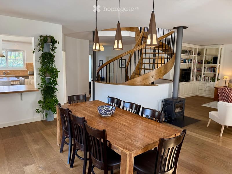Open-plan living and dining area with a wooden dining table, chairs, a bowl, a wooden staircase, a bookshelf, a television, a sofa, and a wood stove.