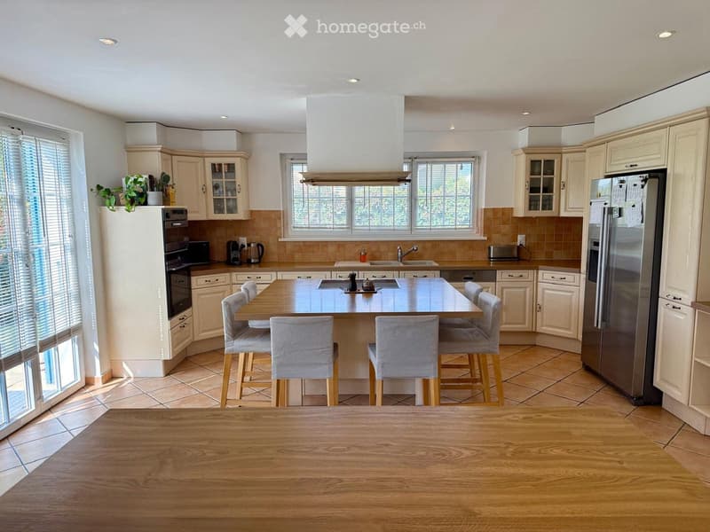 Bright kitchen with cream-colored cabinets, stainless steel appliances, a central island with four stools, and a dining table in the foreground.