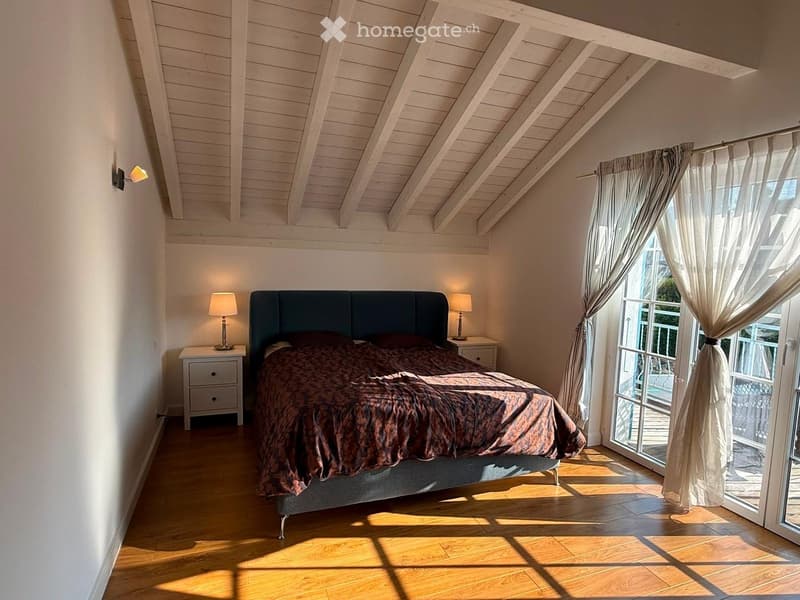 Cozy attic bedroom with wooden slanted ceilings, a large bed with dark headboard, nightstands with lamps, and large windows with sheer curtains leading to a balcony.