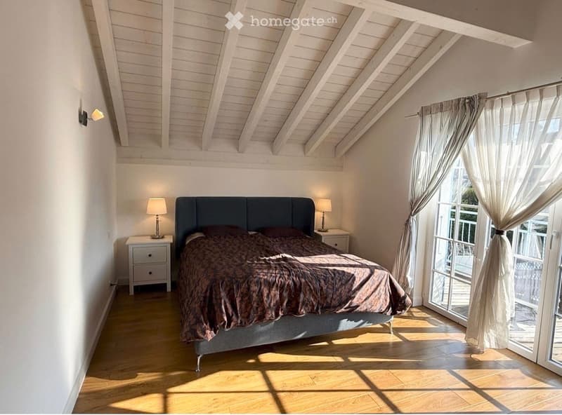 Bright bedroom with wooden flooring, white walls, and a slanted wooden ceiling. Features a dark blue headboard bed with a patterned duvet, two bedside tables with lamps, and large windows with sheer curtains leading to a balcony.