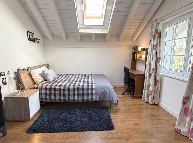 Cozy attic bedroom with wooden slanted ceilings, skylight windows, a bed with plaid bedding, a nightstand, a desk with a chair, and a patterned curtain.