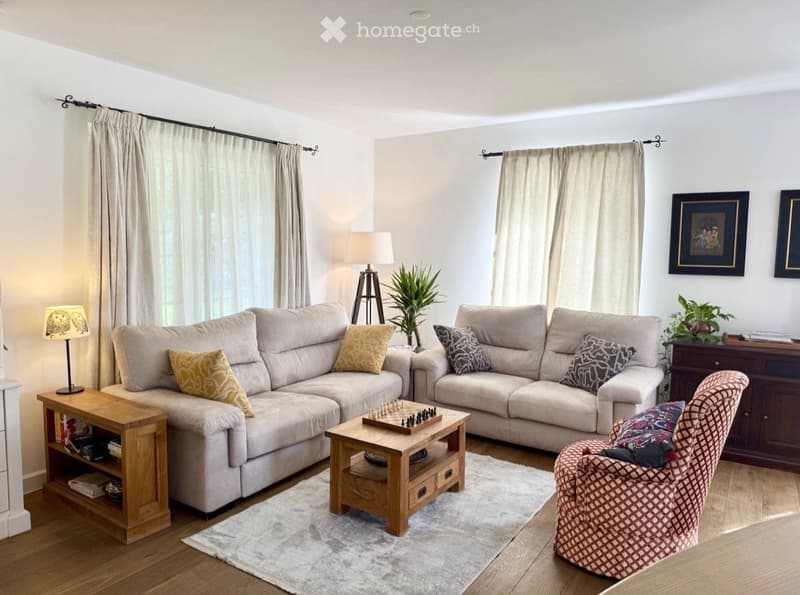 A modern living room with a large sectional sofa, a wooden coffee table, a patterned armchair, and two table lamps. The room features light-colored walls, wooden flooring, and large windows with curtains.