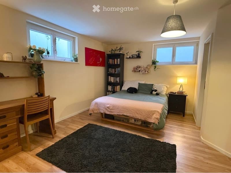Cozy bedroom with a double bed, nightstand, desk, bookshelf, and a large window letting in natural light. The room features a wooden floor, a dark rug, and decorative items including a vase with flowers and a picture on the wall.