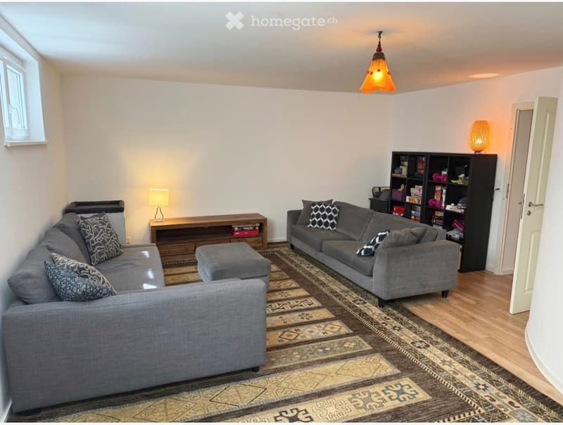 Modern living room with grey sectional sofa, ottoman, wooden coffee table, two table lamps, and a bookshelf with decorative items.