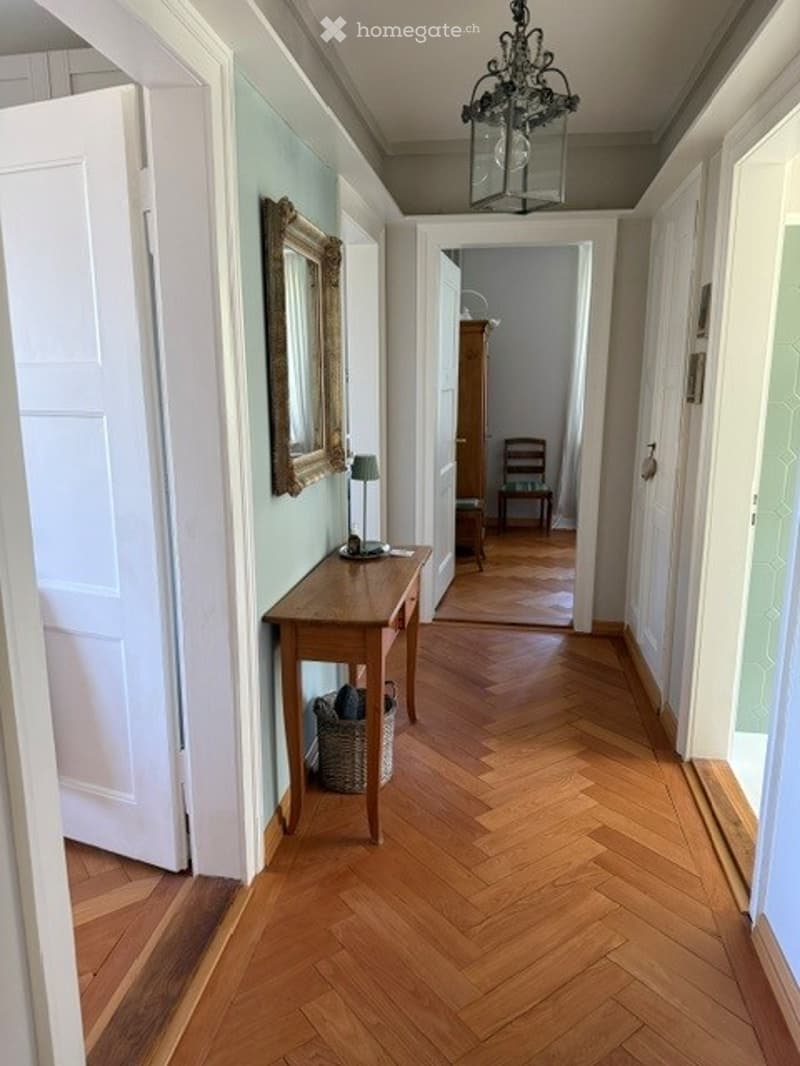 Elegant hallway with herringbone wooden flooring, white walls, and a small wooden console table with a mirror and lamp. A chandelier hangs from the ceiling.