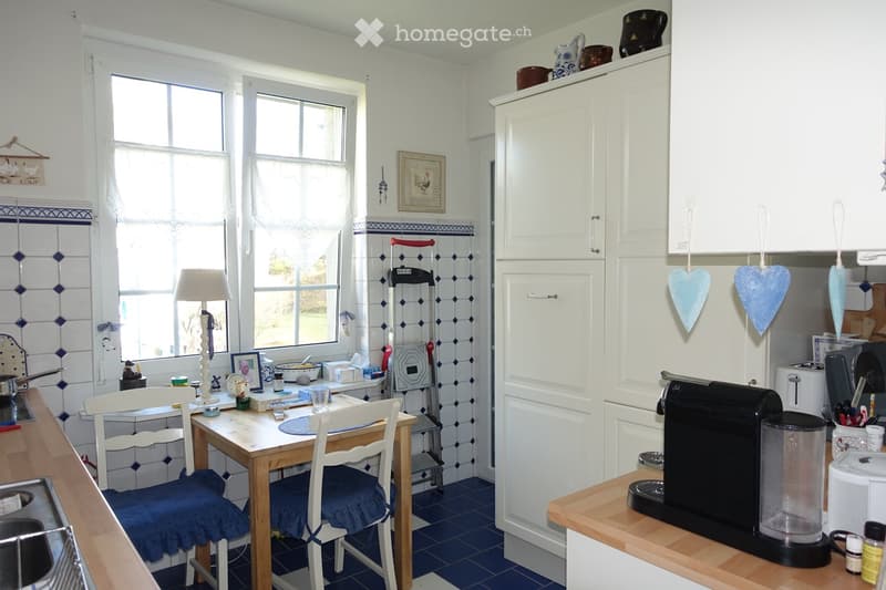 Bright kitchen with white cabinets, blue tiled backsplash, wooden dining table with two chairs, coffee maker, toaster, and various kitchen items on the counter.